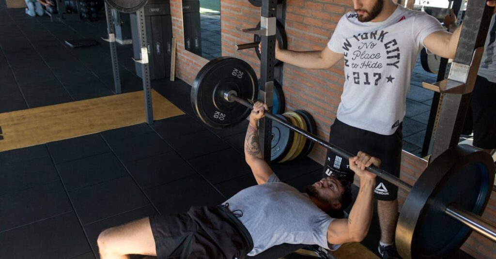 A strong man lifts weights on a bench while a trainer assists in a modern gym setup.