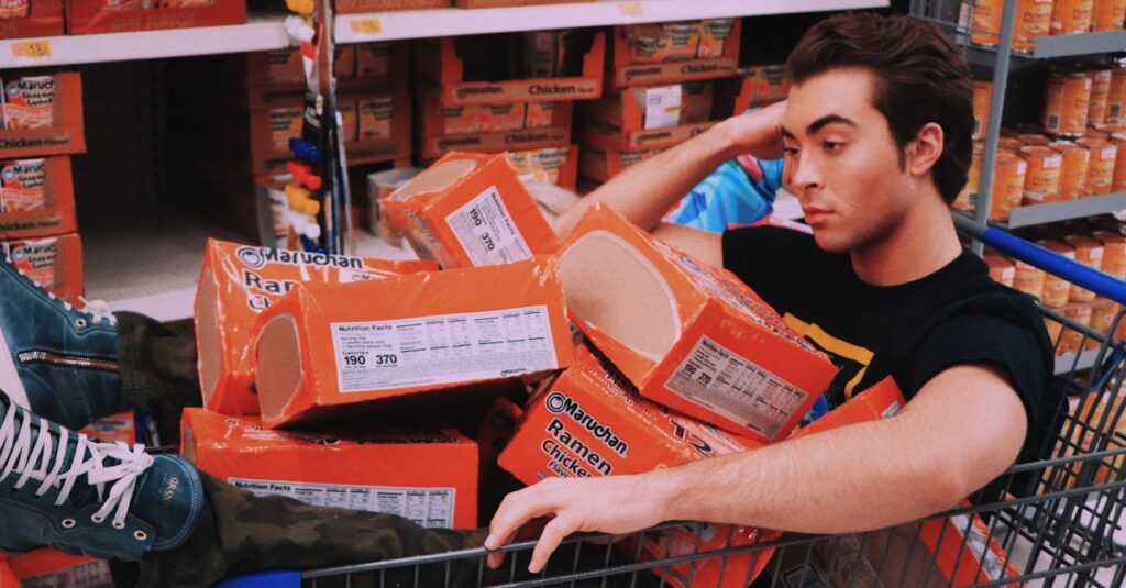 A man lounging in a shopping cart filled with Maruchan ramen in a supermarket aisle.