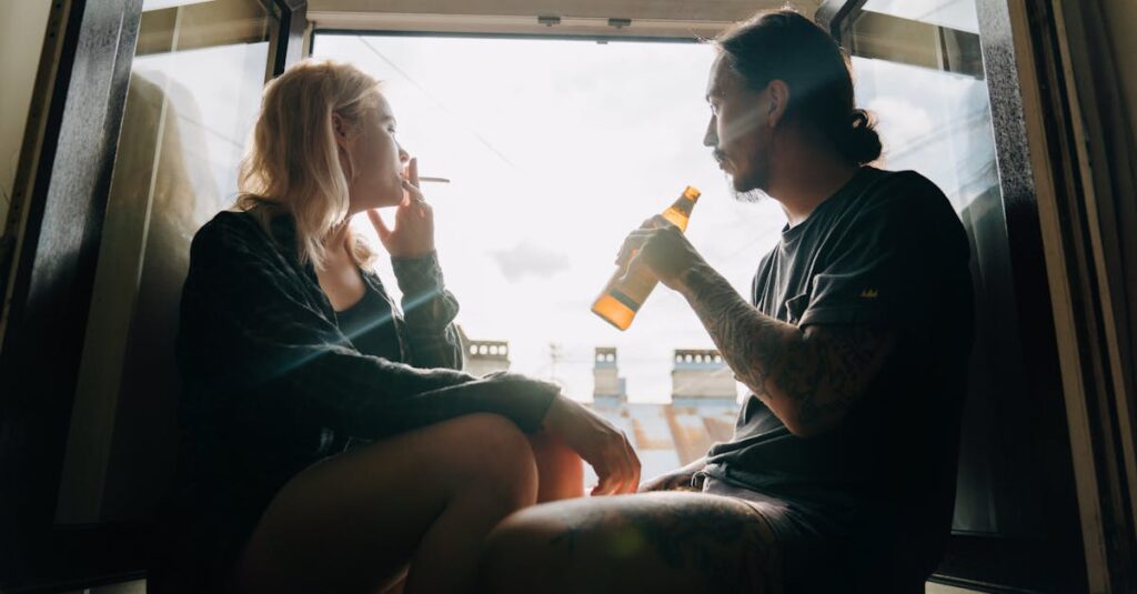 A couple sits by an open window enjoying a beer and cigarette, sharing a quiet moment together.