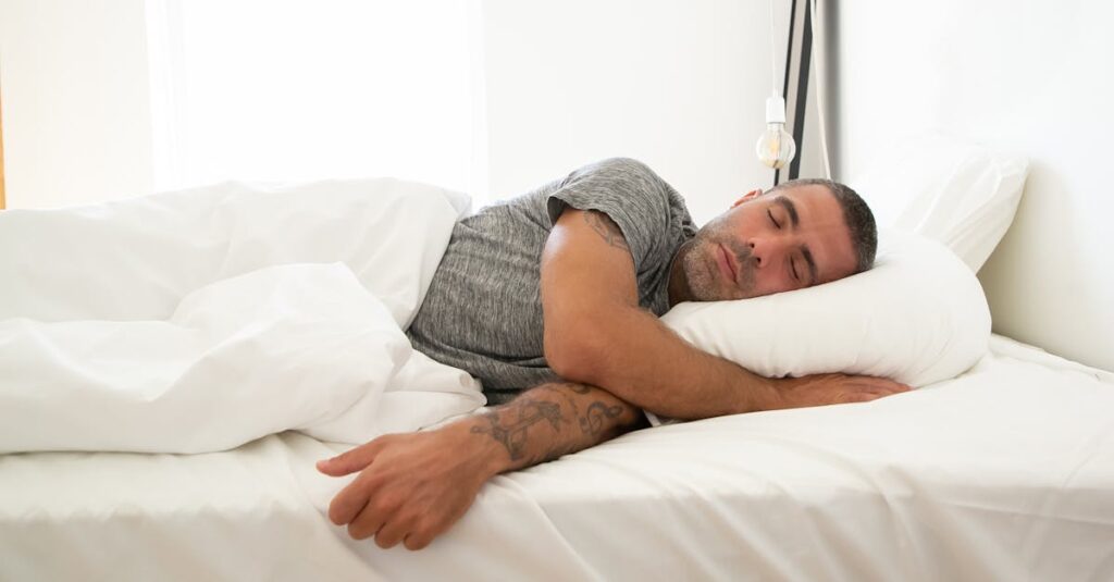 Adult man sleeping soundly on a white bed, tattoo visible, in a bright room.