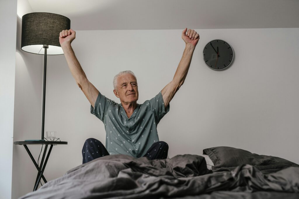 Senior man in pajamas waking up with arms stretched in a cozy bedroom setting.
